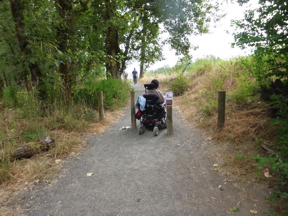 Sandy River Delta Trailhead. A person in a motorized chair is rolling through the gap between two poles that prevent cars from entering the trail. The ground is compacted gravel – encroaching grass on hard-packed soil surface.