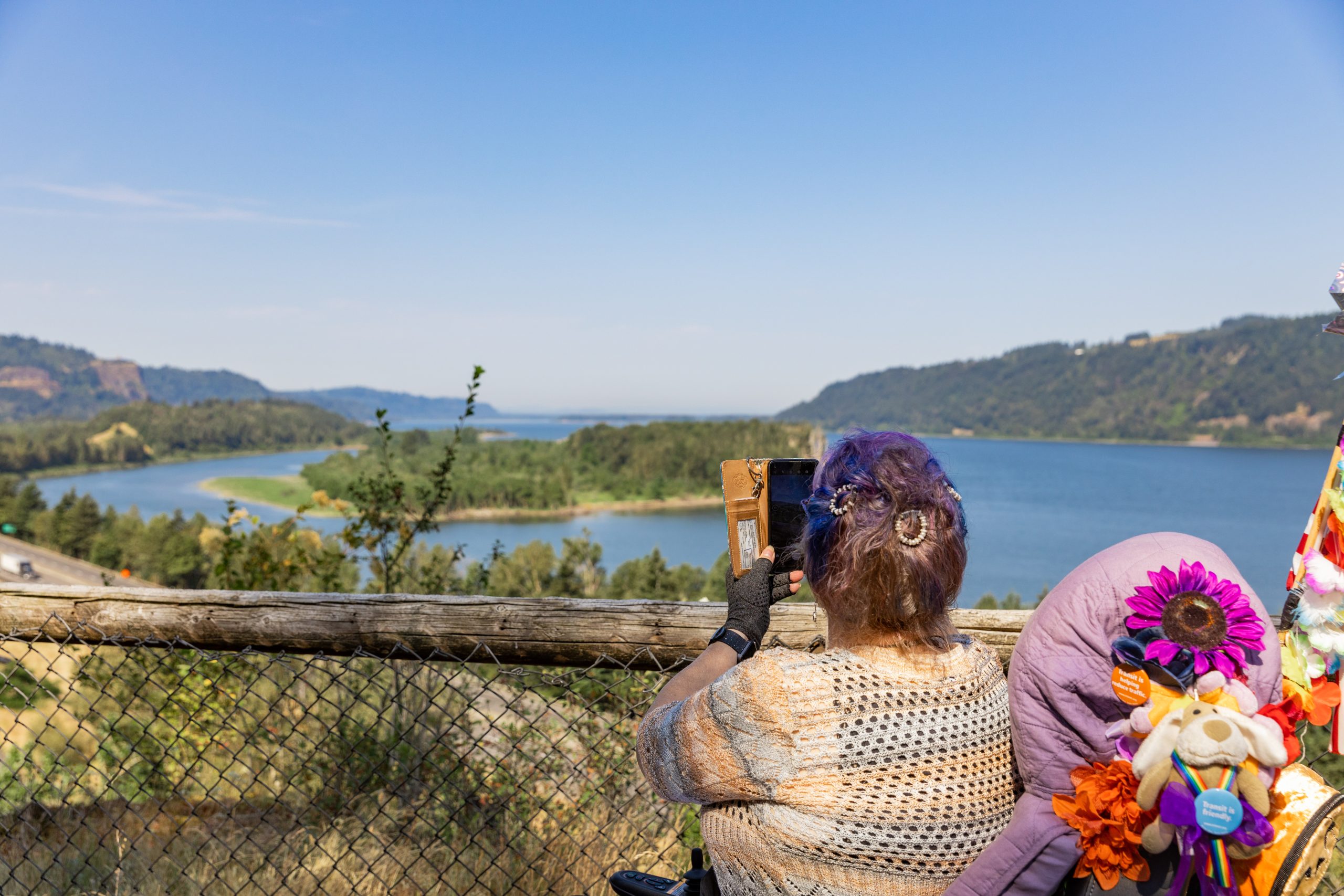 The picture shows a view from the Overlook Trail with the Columbia River taking up most of the photo. The shore on either side is visible with an island sitting in the middle. In the foreground a woman in a motorized wheelchair is taking a picture with her phone over a fence.